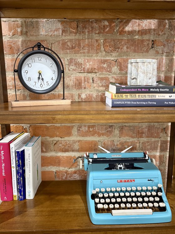A shelf in the office holding a clock, a teal typewriter, and books on trauma and healing