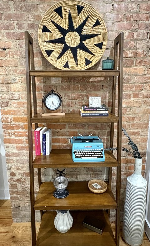 The bookshelf against exposed brick, holding a woven basket, a clock, books, and a teal typewriter